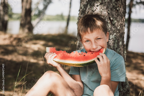 Happy teenager boy eating watermelon outdoors. Summer food and healthy snack. Portrait of cute joyful child sitting on green grass and enjoying tasty juicy sweet fruit
