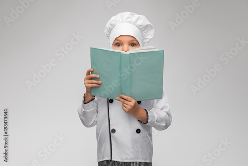 cooking, culinary and profession concept - little girl in chef's toque and jacket reading cook book over grey background