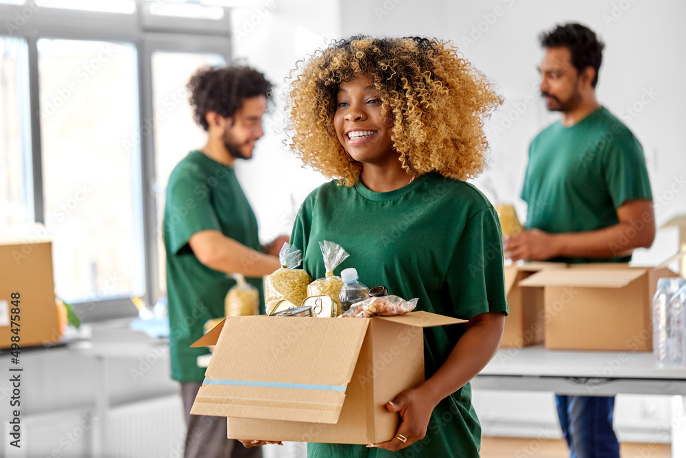 © Syda Productions - charity, donation and volunteering concept - happy smiling female volunteer with food in box and international group of people at distribution or refugee assistance center