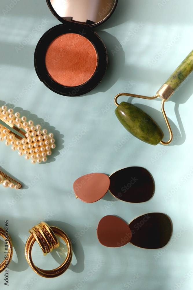 Coral blush, jade roller, pearl barrettes and gold earrings on bright blue background. Flat lay. Coral blush, jade roller, pearl barrettes and gold earrings on bright blue background. Flat lay.