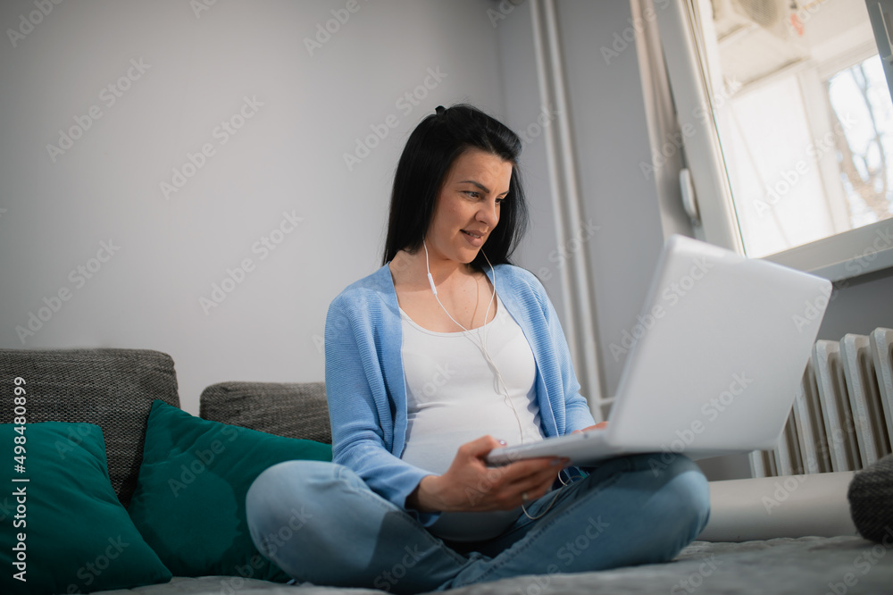 Nice-looking caucasian pregnant female looks at monitor of white laptop and type on keyboard with her left hand. Woman sits on bed with crossed legs and smiles. She is black-haired and wears earphones Nice-looking caucasian pregnant female looks at monitor of white laptop and type on keyboard with her left hand. Woman sits on bed with crossed legs and smiles. She is black-haired and wears earphones