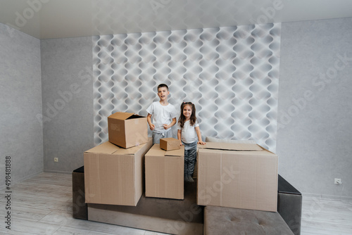 Brother and sister are playing and enjoying moving to a new apartment against the background of cardboard boxes on the couch. Purchase of real estate. Housewarming, delivery. © Andrii