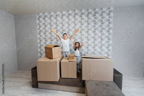 Brother and sister are playing and enjoying moving to a new apartment against the background of cardboard boxes on the couch. Purchase of real estate. Housewarming, delivery.