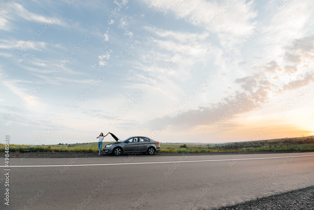 A young girl stands near a broken-down car in the middle of the highway during sunset and tries to call for help on the phone. Waiting for help. Car service. Car breakdown on road. A young girl stands near a broken-down car in the middle of the highway during sunset and tries to call for help on the phone. Waiting for help. Car service. Car breakdown on road.