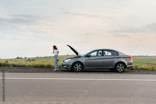 A young girl stands near a broken car in the middle of the highway during sunset and tries to call for help on the phone and start the car. Waiting for help. Car service. Car breakdown on the road. © Andrii