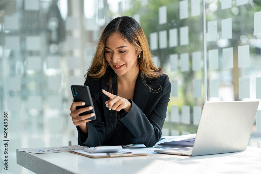 Young beautiful asian woman using smartphone and working with laptop while sitting at office desk, working from home concept.