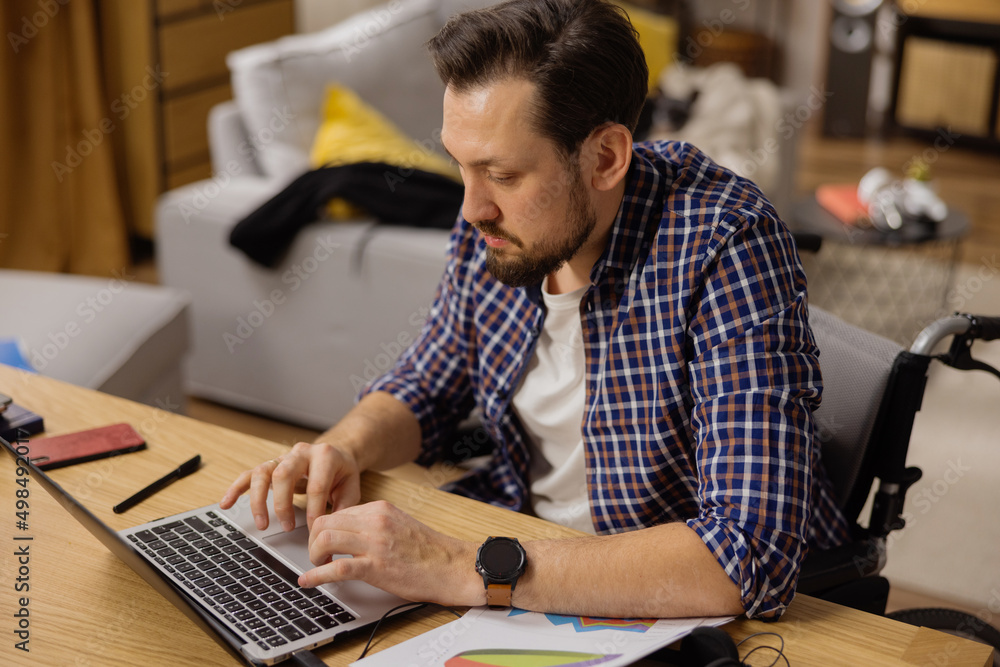 Portrait of a bearded man sitting in a wheelchair working on a laptop. A middle-aged boy is preparing presentations based on data lying next to him. Work at home. Portrait of a bearded man sitting in a wheelchair working on a laptop. A middle-aged boy is preparing presentations based on data lying next to him. Work at home.