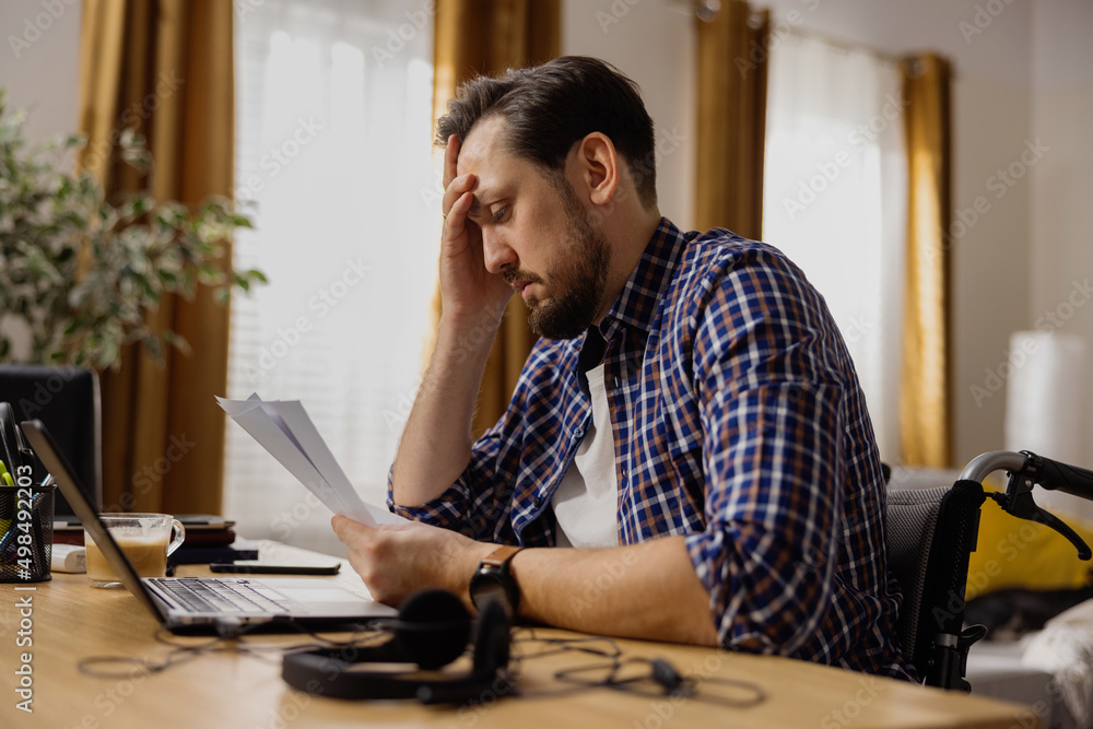 A man with a disability wearing a navy blue shirt sits alone in his office while working . The focused bearded man props his head up, staring at the documents lying before him. A man with a disability wearing a navy blue shirt sits alone in his office while working . The focused bearded man props his head up, staring at the documents lying before him.