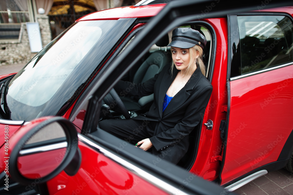 Portrait of beautiful blonde sexy fashion woman model in cap and in all black with bright makeup sit and drive red city car.
