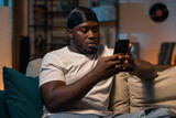 Evening in the living room.A man is relaxing on the sofa dressed in jeans and a white T-shirt. A student holds phone in hands sends messages, chats with his girlfriend, writes an email.