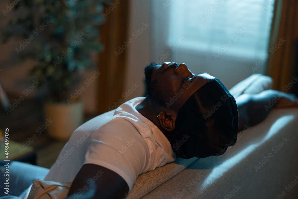 Man wearing black bandana is resting on couch with head tilted back. Men  with stubble is experiencing negative emotions, breakdown, sad day, dark  night lantern light from outside window. Stock Photo
