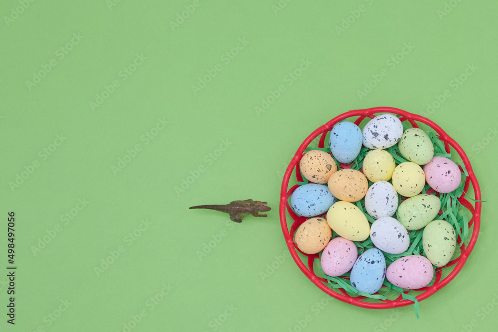 A basket with eggs in various colors and a dinosaur walking towards them on a green background with copy space. Minimal Easter scene. A basket with eggs in various colors and a dinosaur walking towards them on a green background with copy space. Minimal Easter scene.
