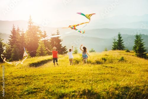 Three children running towards sun in mountains at sunset. Kids play with kites. Happy summer vacation and healthy lifestyle concept. Sunlit mountain ranges with coniferous trees.