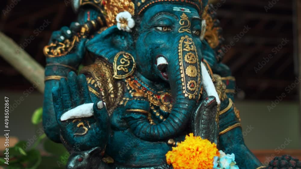 An old stone statue of Ganesha covered with moss with a wreath of yellow flowers on his neck standing on the street with green plants An old stone statue of Ganesha covered with moss with a wreath of yellow flowers on his neck standing on the street with green plants