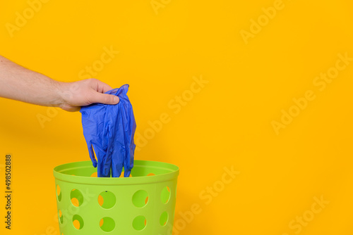 Medical gloves are thrown into a green trash can for disposal and recycling, yellow background, side view, copy space.