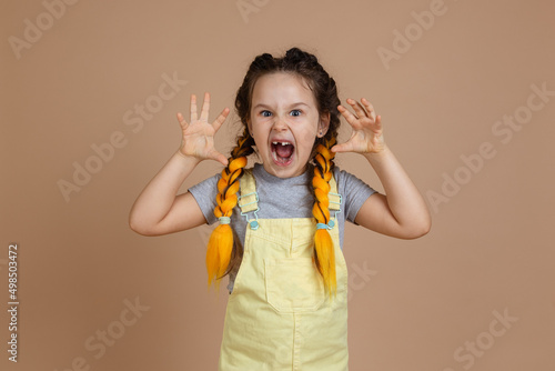 Portrait of playful little girl with yellow kanekalon pigtails, pretending to be scary frightening with hands and opened mouth wearing yellow jumpsuit and gray t-shirt on beige background.