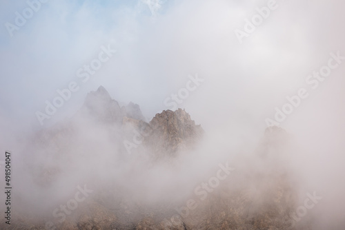 Scenic alpine landscape with rocky mountains in dense low clouds in morning sunlight. Colorful mountain scenery with sharp rocks among thick low clouds. Awesome view to high rockies in low cloudiness. © Daniil
