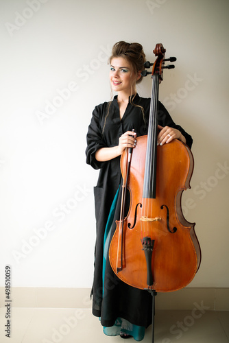 a girl stands with a cello against a white wall, a full-length portrait of a female cellist