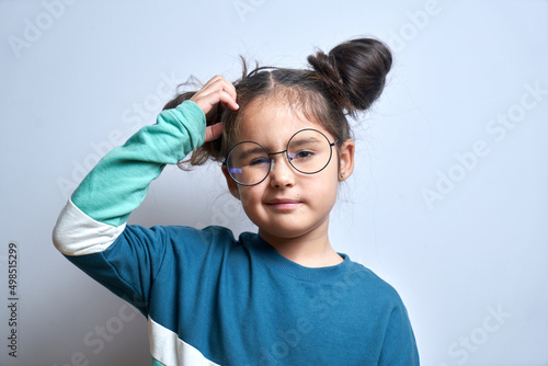 Funny little girl in glasses scratching her head doubts, makes decision isolated on yellow studio background