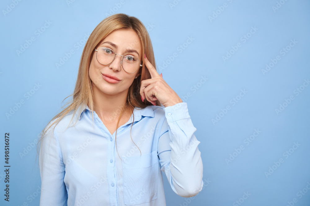 Smart blonde girl in business shirt and glasses touches her head thinks doubts, makes decision isolated on blue studio background Smart blonde girl in business shirt and glasses touches her head thinks doubts, makes decision isolated on blue studio background