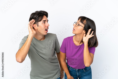 Young couple isolated on isolated white background listening to something by putting hand on the ear
