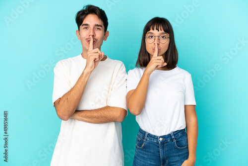 Young mixed race couple isolated on blue background showing a sign of closing mouth and silence gesture