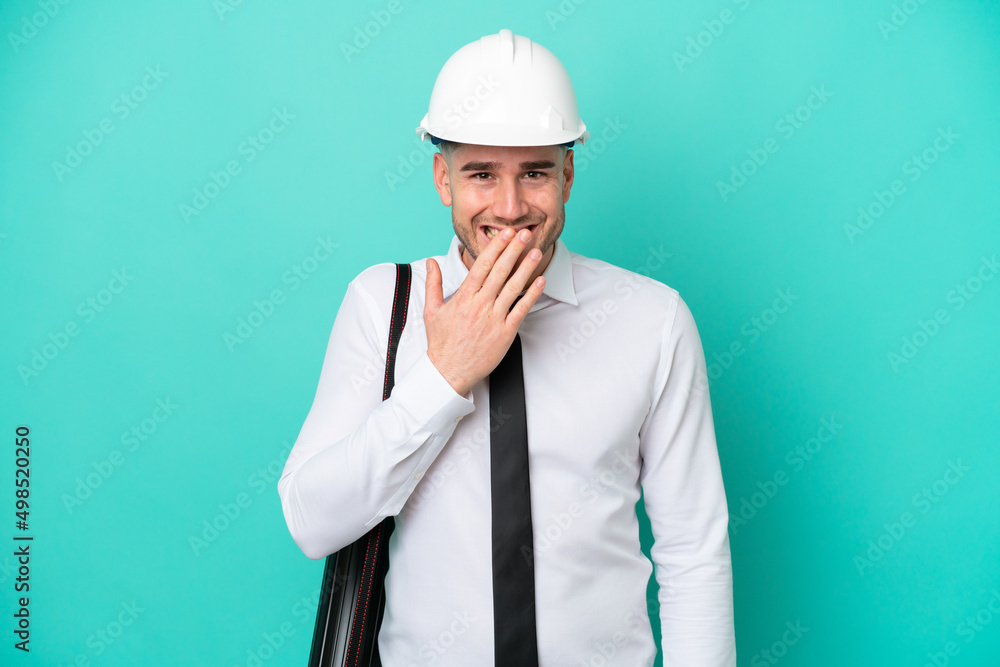 Young architect caucasian man with helmet and holding blueprints isolated on blue background happy and smiling covering mouth with hand