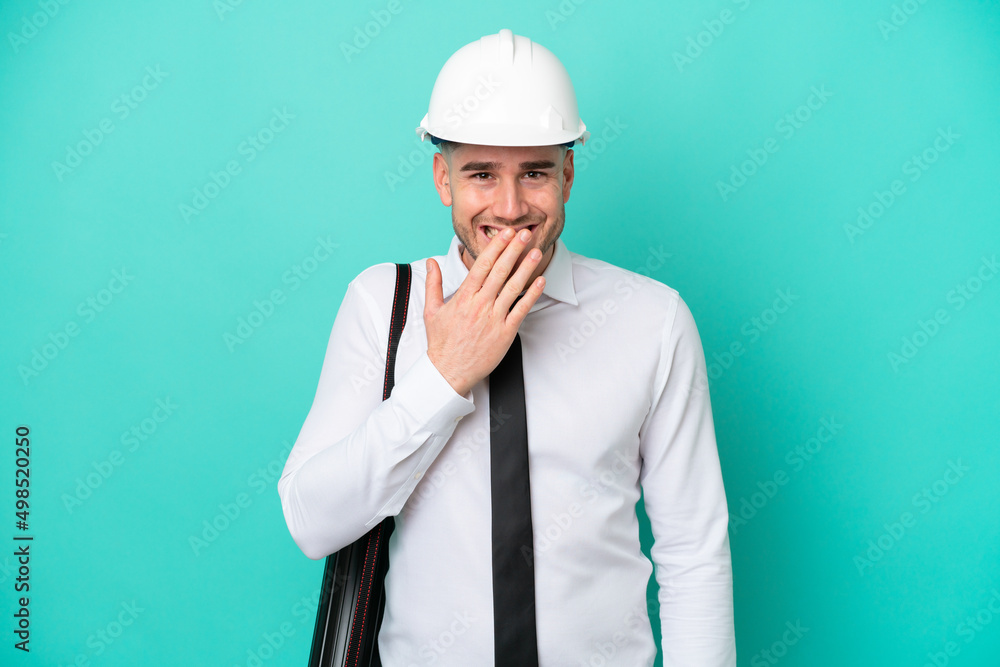 Young architect caucasian man with helmet and holding blueprints isolated on blue background happy and smiling covering mouth with hand Young architect caucasian man with helmet and holding blueprints isolated on blue background happy and smiling covering mouth with hand