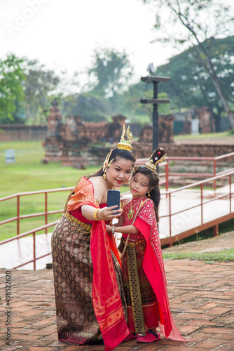 An Asian woman in a red Thai traditional dress take a picture with her daughter by her phone while traveling in Ayutthaya