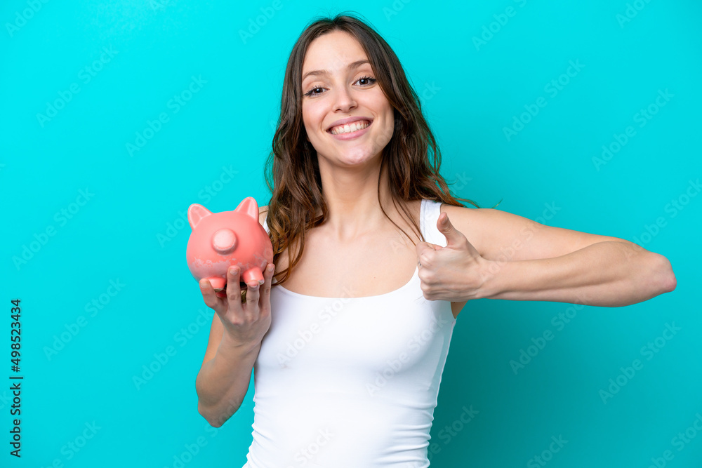 Young Caucasian woman holding a piggybank isolated on blue bakcground with thumbs up because something good has happened