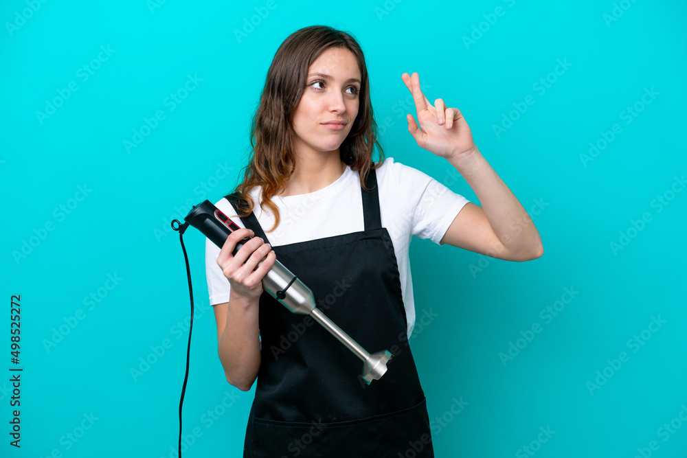 Young caucasian cooker woman using hand blender isolated on blue background with fingers crossing and wishing the best Young caucasian cooker woman using hand blender isolated on blue background with fingers crossing and wishing the best