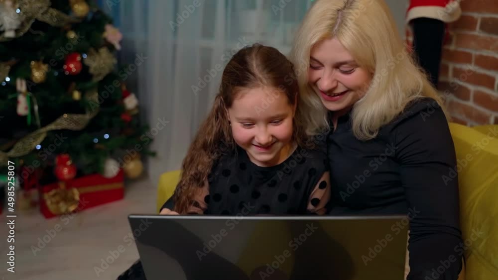 Females persons sitting on sofa and using laptop at home indoors. Mother and daughter having fun online, they smile and laugh. 30s woman and young girl looking notebook screen. Xmas decor in room Females persons sitting on sofa and using laptop at home indoors. Mother and daughter having fun online, they smile and laugh. 30s woman and young girl looking notebook screen. Xmas decor in room