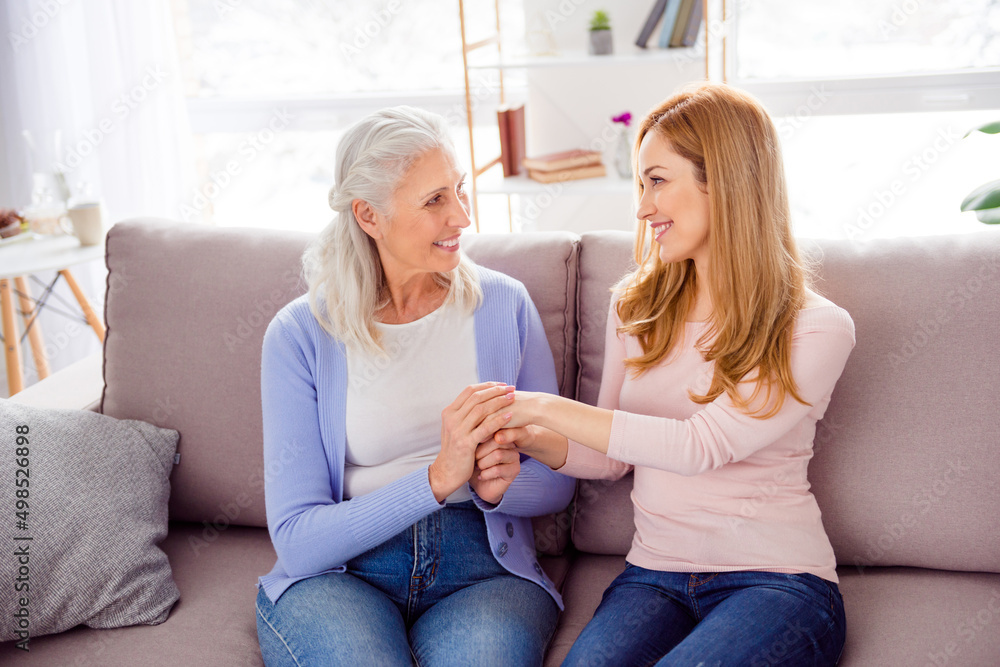 Photo of two cheerful charming lady sitting hold arms look each other house indoors Photo of two cheerful charming lady sitting hold arms look each other house indoors