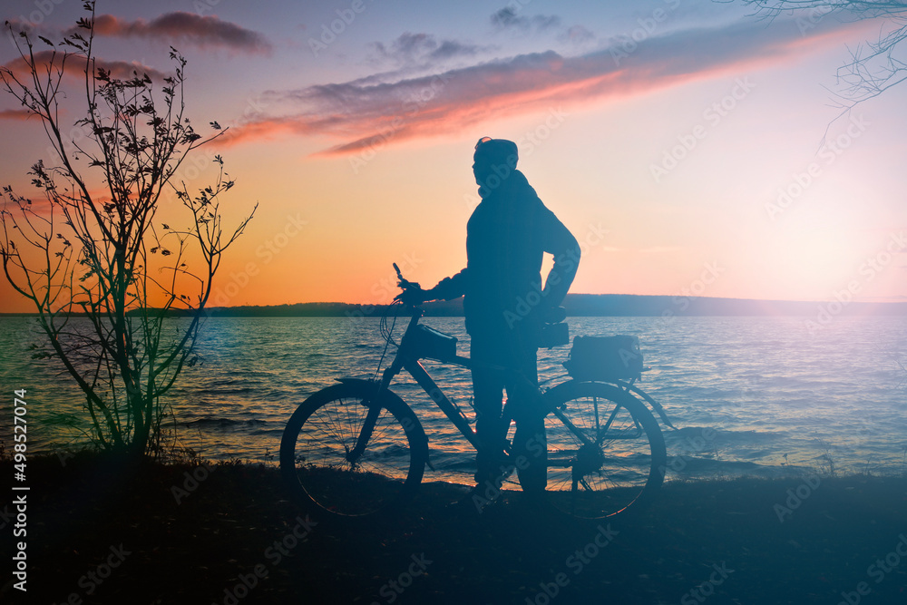 Obraz premium Silhouette of a female cyclist and a bicycle against the backdrop of a sunset over a lake. Sports action.