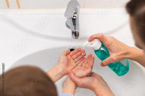 A young mother squeezes liquid soap from a dispenser onto her child's hands for washing hands at home over the washbasin in the bathroom. Personal hygiene. Selective focus. Close-up