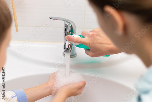 A young mother squeezes liquid soap from a dispenser onto her child s hands for washing hands at home over the washbasin in the bathroom. Personal hygiene. Selective focus. Close-up