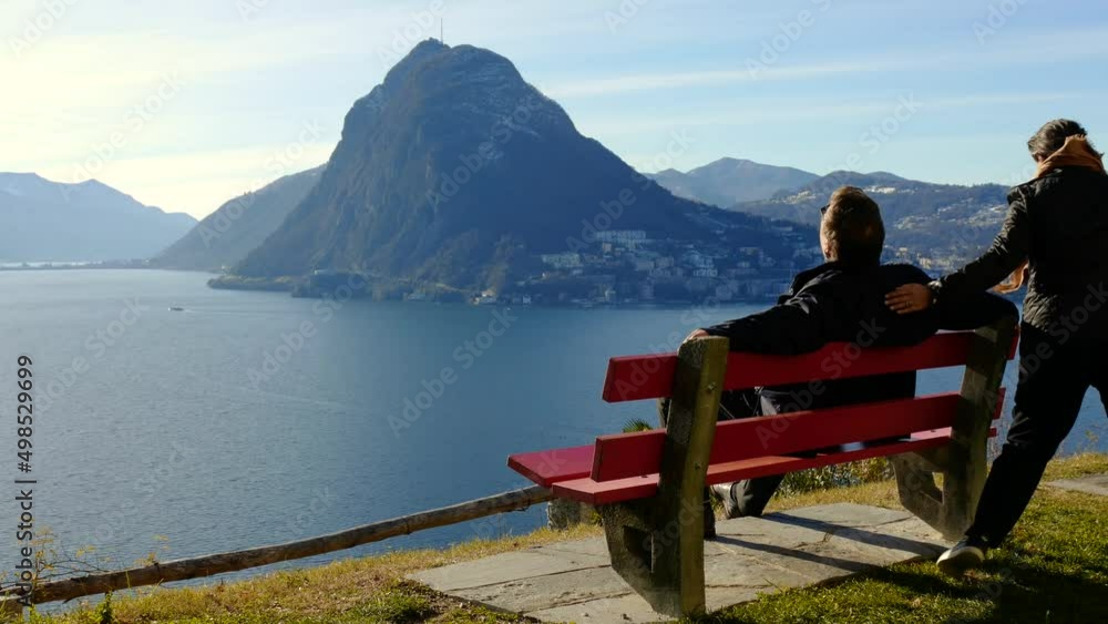 Couple Sitting and Hugging on a Bench with Lake Lugano and Mountain View in a Sunny Day and Cityscape in Lugano, Ticino, Switzerland. Couple Sitting and Hugging on a Bench with Lake Lugano and Mountain View in a Sunny Day and Cityscape in Lugano, Ticino, Switzerland.