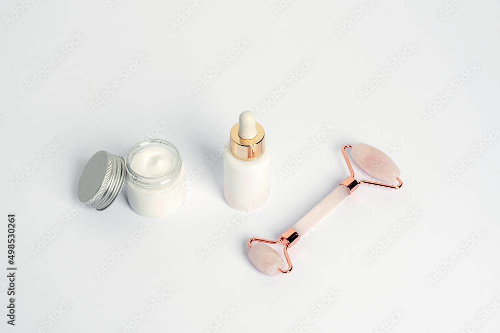 Cream in a jar, serum bottle and crystal rose quartz facial roller on white background, top view