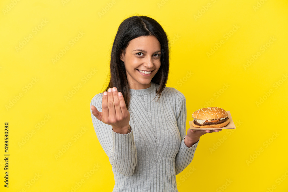 Young caucasian woman holding a burger isolated on yellow background inviting to come with hand. Happy that you came Young caucasian woman holding a burger isolated on yellow background inviting to come with hand. Happy that you came