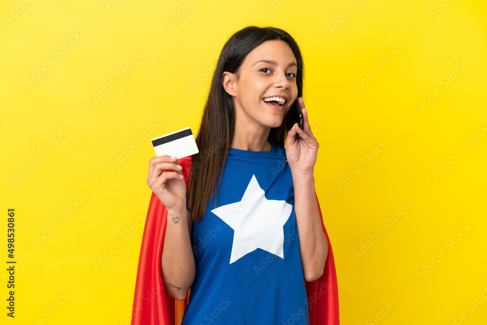 Super Hero woman isolated on yellow background keeping a conversation with the mobile phone and holding a credit card Super Hero woman isolated on yellow background keeping a conversation with the mobile phone and holding a credit card
