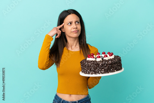 Young caucasian woman holding birthday cake isolated on blue background having doubts and thinking