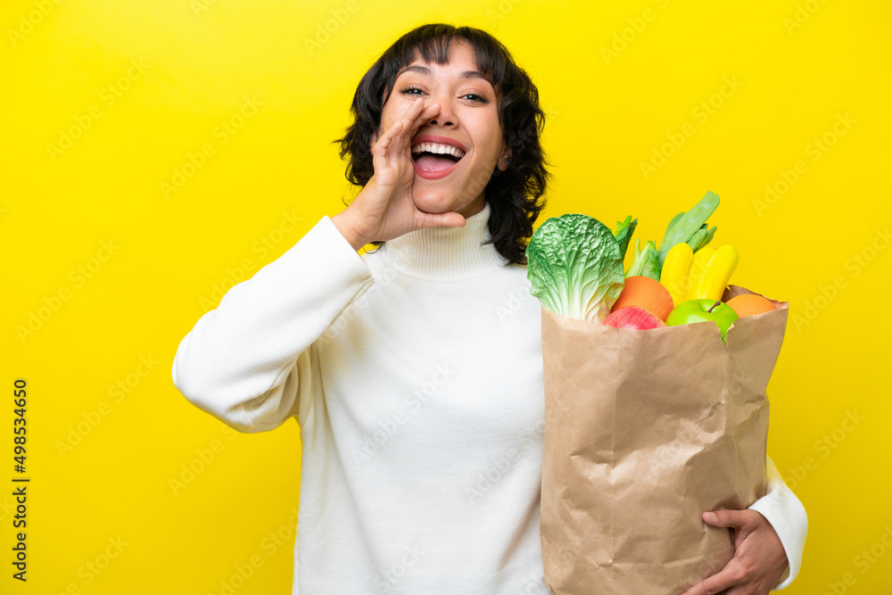 Young Argentinian woman holding a grocery shopping bag isolated on yellow background shouting with mouth wide open Young Argentinian woman holding a grocery shopping bag isolated on yellow background shouting with mouth wide open