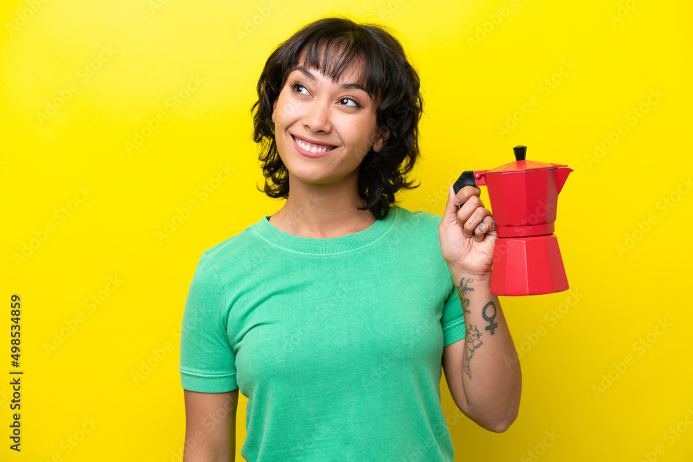 Young Argentinian woman holding coffee pot isolated on yellow background looking up while smiling Young Argentinian woman holding coffee pot isolated on yellow background looking up while smiling