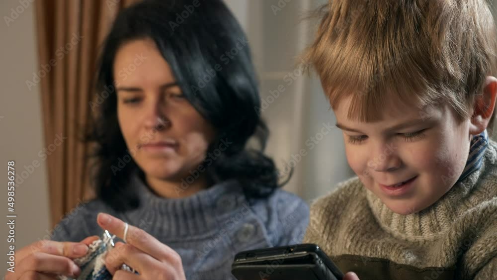 Child Kid Boy Use Watching Smartphone Internet Social Media. Mother Knit with Knitting Needles near Window. Cold Winter Season. 2x Slow motion 60fps 4K Child Kid Boy Use Watching Smartphone Internet Social Media. Mother Knit with Knitting Needles near Window. Cold Winter Season. 2x Slow motion 60fps 4K