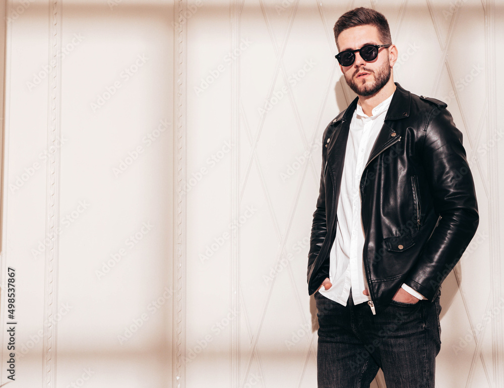 Handsome confident model. Sexy stylish man in biker leather jacket and black jeans. Fashion hipster male posing near white wall in studio interior at sunny day. Shadow from window in sunglasses Handsome confident model. Sexy stylish man in biker leather jacket and black jeans. Fashion hipster male posing near white wall in studio interior at sunny day. Shadow from window in sunglasses