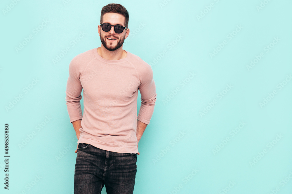 Portrait of handsome smiling model. Sexy stylish man dressed in pink sweater and jeans. Fashion hipster male posing near blue wall in studio. In sunglasses. Isolated Portrait of handsome smiling model. Sexy stylish man dressed in pink sweater and jeans. Fashion hipster male posing near blue wall in studio. In sunglasses. Isolated