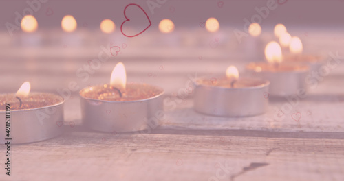 Image of hearts falling over lit tea candles on wooden surface in background