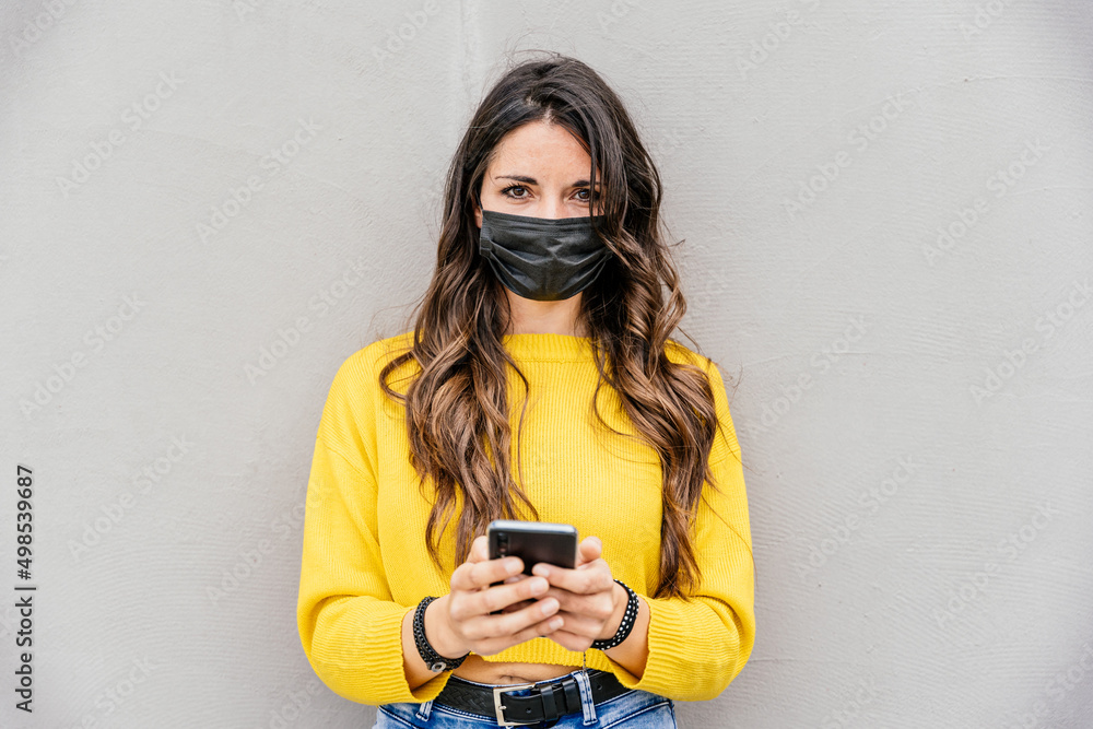 Portrait of a young beautiful woman with protective face mask during the global Coronavirus Covid-19 pandemic while using the smartphone device - Friendly female on a gray wall background - Copy space