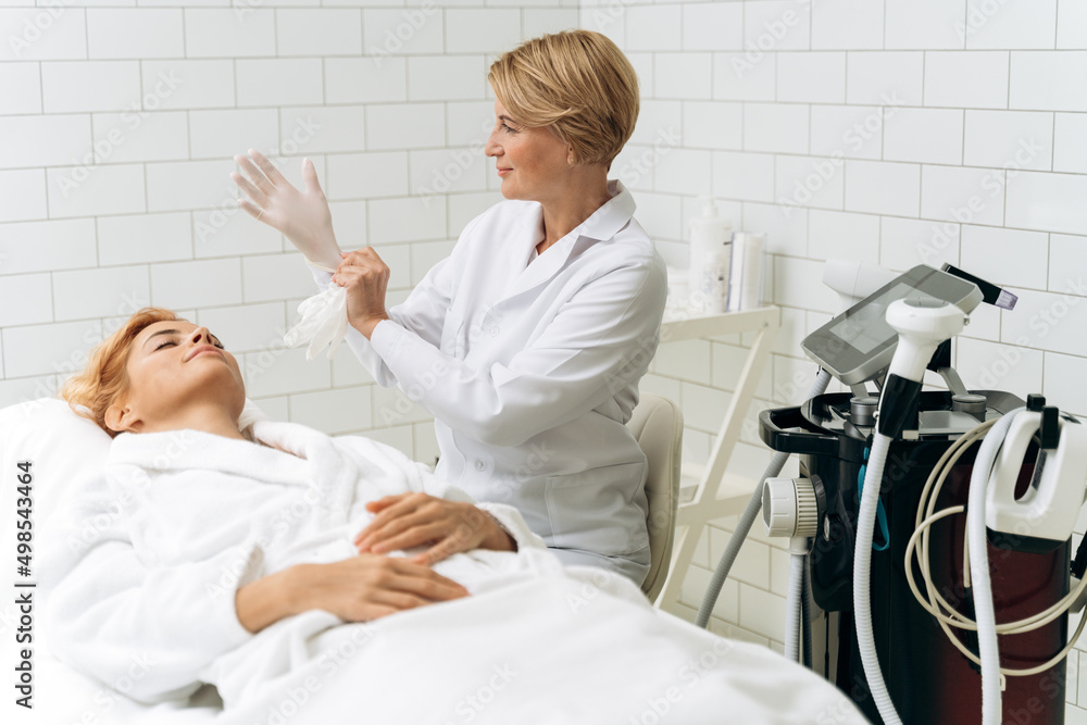 Charming senior beautician in white lab coat taking on protective gloves with smile Charming senior beautician in white lab coat taking on protective gloves with smile
