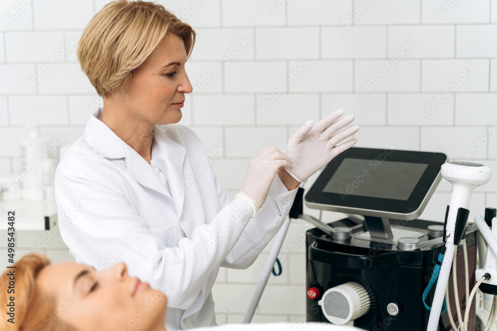 Charming senior beautician in white lab coat taking on protective gloves with smile Charming senior beautician in white lab coat taking on protective gloves with smile